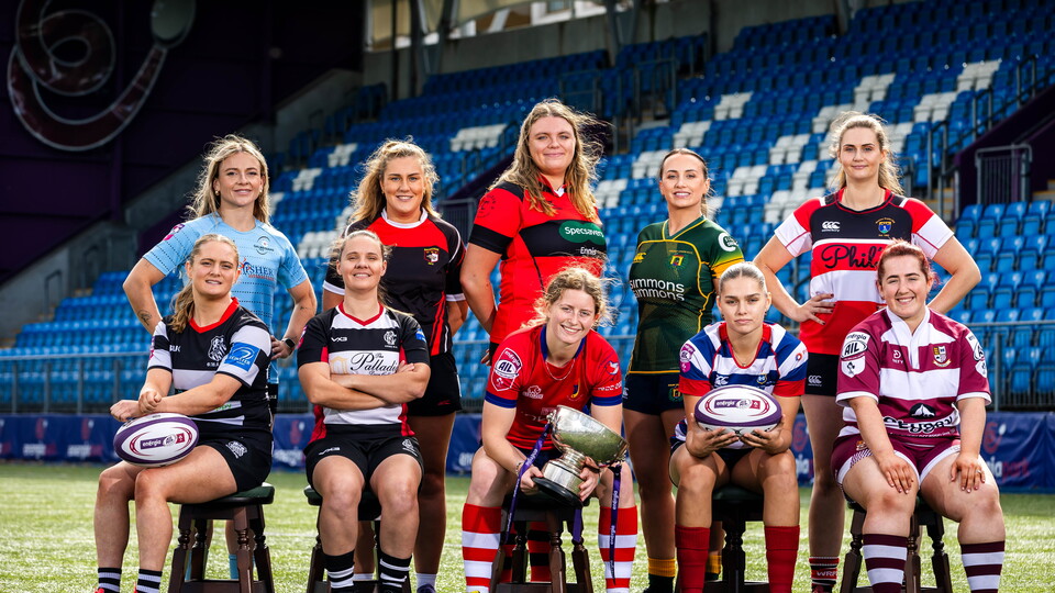 Eleven women rugby players in a variety of team kits, sitting on stools, pose for a photograph on a pitch.
