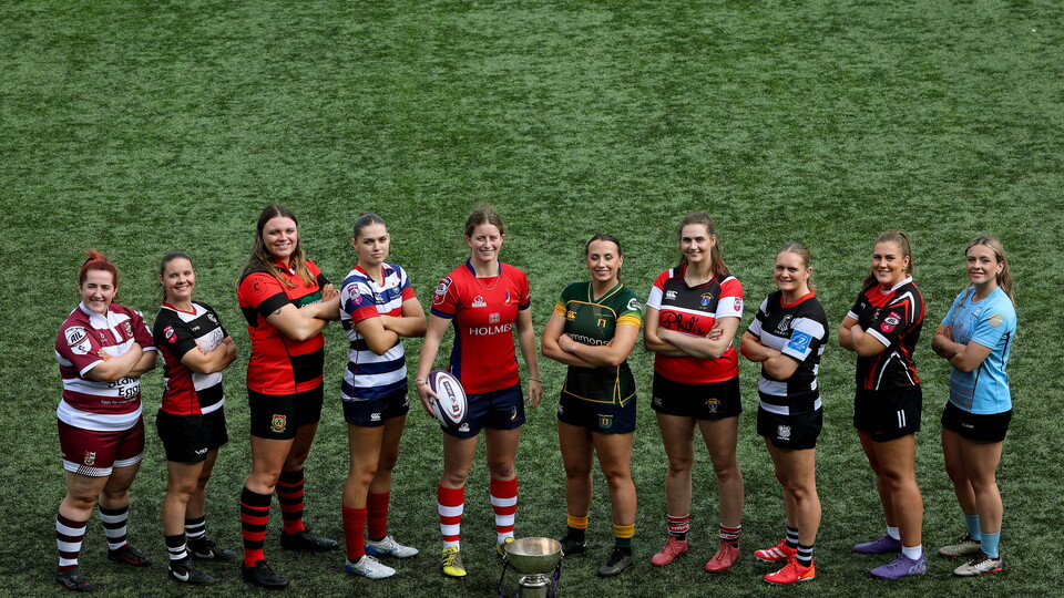 Eleven female rugby players in team kits line up on a rugby pitch with a trophy